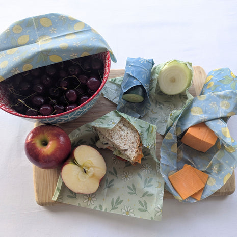 Reusable beeswax food wraps shown in everyday kitchen use with a covered bowl of cherries, wrapped cucumber and onion, cheddar cheese, a sandwich, and a cut red apple.  The natural wraps help cover bowls, protect produce, store cheese, and pack lunches while offering a practical reusable alternative to disposable plastic wrap.