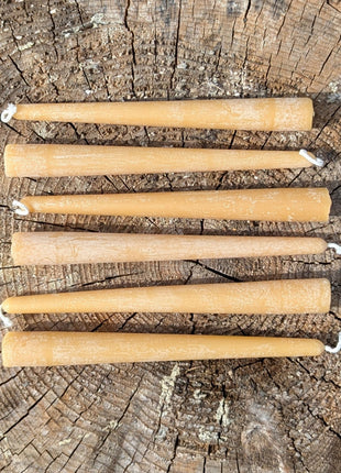 Overhead photograph of six natural beeswax taper candles arranged across the growth rings of a weathered tree stump.  The rustic outdoor setting clearly shows the complete six-candle quantity, warm beeswax colour, individual cotton wicks and natural variations between the candles.