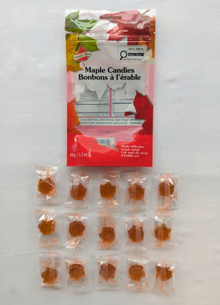 A 90 g package of Canadian maple candies is displayed above approximately fifteen individually wrapped maple-leaf-shaped candies on a white background.  The image gives customers a clear sense of the candy size, amber colour, wrapping, and approximate quantity contained in the bag.