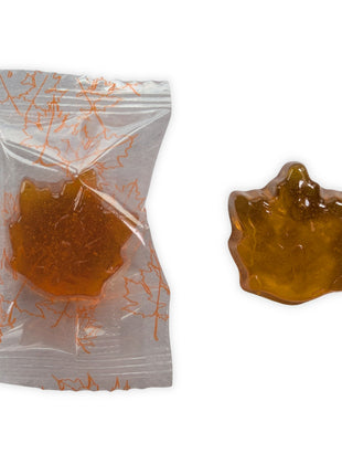 A close-up of one individually wrapped maple-leaf hard candy beside an unwrapped candy on a clean white background.  The image shows the transparent wrapper printed with orange maple leaves as well as the candy’s detailed leaf shape, glossy surface, golden amber colour, and translucent appearance.