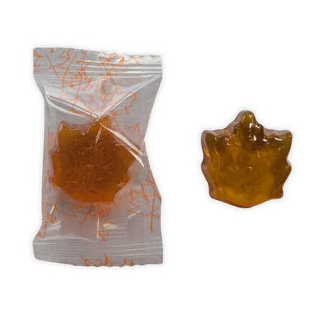A close-up of one individually wrapped maple-leaf hard candy beside an unwrapped candy on a clean white background.  The image shows the transparent wrapper printed with orange maple leaves as well as the candy’s detailed leaf shape, glossy surface, golden amber colour, and translucent appearance.
