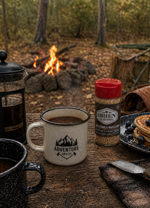 Maple sugar shaker beside camp coffee, pancakes, and a French press on a rustic table near a forest campfire.
