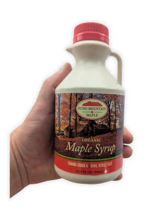 A hand holding the 500 mL Echo Mountain organic maple syrup jug against a clean white background to show its true size and proportions.  The front label, red screw cap, pouring spout, handle and Canada Grade A dark, robust taste designation are clearly visible.