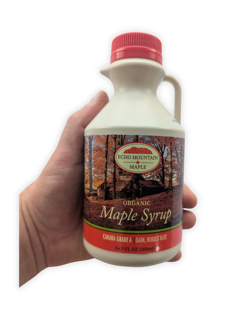 A hand holding the 500 mL Echo Mountain organic maple syrup jug against a clean white background to show its true size and proportions.  The front label, red screw cap, pouring spout, handle and Canada Grade A dark, robust taste designation are clearly visible.