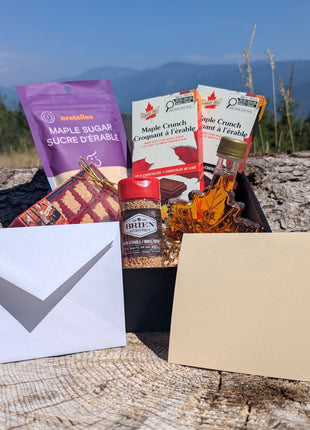 LOONMOON Premium Maple Gift Box displayed outdoors against a British Columbia mountain backdrop with Canadian maple syrup, soft maple candies, maple sugar, and two maple chocolate bars arranged in a black gift box.  A blank folded greeting card and matching envelope are shown in front, highlighting the complete gift-ready presentation.