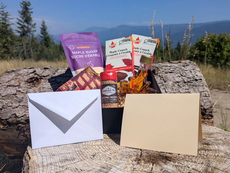 LOONMOON Premium Maple Gift Box displayed outdoors against a British Columbia mountain backdrop with Canadian maple syrup, soft maple candies, maple sugar, and two maple chocolate bars arranged in a black gift box.  A blank folded greeting card and matching envelope are shown in front, highlighting the complete gift-ready presentation.