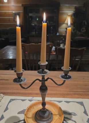 Three beeswax taper candles with subtle flickering flames, gently rotating from left to right in a warm rustic, homestead setting.