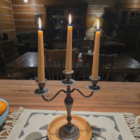 Three beeswax taper candles with subtle flickering flames, gently rotating from left to right in a warm rustic, homestead setting.