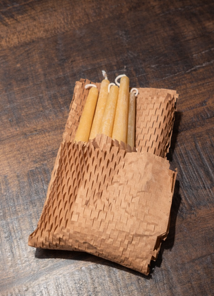 “Six beeswax taper candles partially wrapped in honeycomb kraft packing paper on a wooden table.