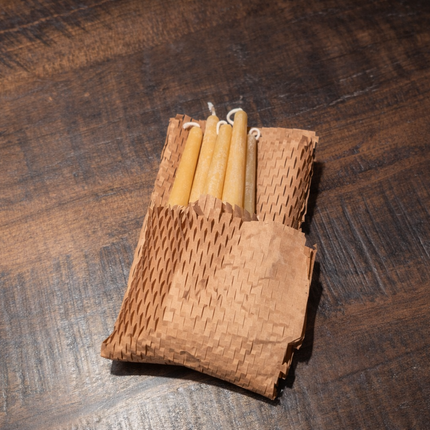 “Six beeswax taper candles partially wrapped in honeycomb kraft packing paper on a wooden table.