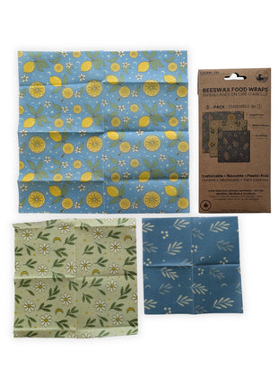 Three reusable beeswax food wraps are laid flat beside their retail package on a clean white background.  The image clearly shows the large lemon-pattern wrap, medium daisy-pattern wrap, and small blue botanical wrap included in the 3-pack.