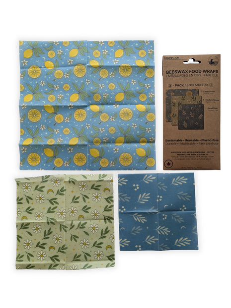 Three reusable beeswax food wraps are laid flat beside their retail package on a clean white background.  The image clearly shows the large lemon-pattern wrap, medium daisy-pattern wrap, and small blue botanical wrap included in the 3-pack.