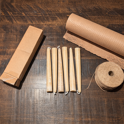 Six beeswax taper candles with visible wax bloom, shown with a kraft box, honeycomb paper wrap, and twine on a dark wood table.