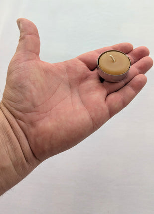 A single beeswax tealight candle rests in an open hand against a clean white background, providing a clear real-world sense of its compact size.  The close view shows the warm natural wax, cotton wick, rounded top, and shallow aluminum tealight cup.