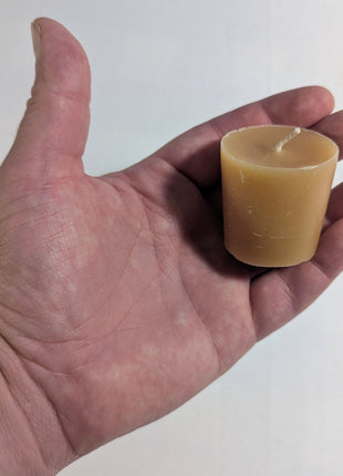 A single honey-gold beeswax votive candle resting in an open hand against a plain white background, providing a clear real-world sense of the candle’s compact size, tapered form, wax texture, and cotton wick.