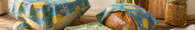 Reusable beeswax food wraps covering a bowl, bread, cheese, and lemon in a rustic kitchen scene with LOONMOON lemon, olive, and blue herbarium wrap patterns.